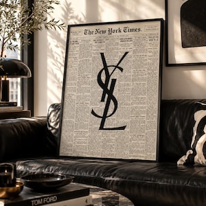 Può includere: Una stampa artistica da parete incorniciata che presenta una pagina di giornale in stile vintage del New York Times con un grande e audace logo YSL nero sovrapposto al centro. La stampa è racchiusa in una sottile cornice nera con colonne di testo.