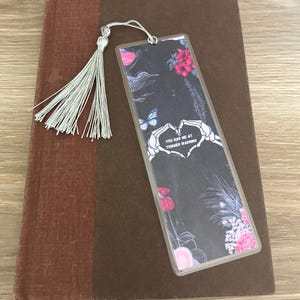 Puede incluir: Un marcapáginas en blanco y negro con un diseño floral y una mano de esqueleto haciendo un corazón. El texto en el marcapáginas dice "You had me at trigger warning".