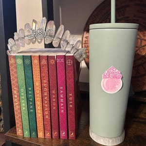 May include: A crystal quartz crown with silver moon accents sits atop a stack of colorful books. A sage green tumbler with a pink sticker and a straw rests on a white stone base. The books have titles like "Imbolc" and "Yule".