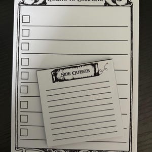 Peut inclure: Un bloc-notes rectangulaire blanc avec une encre noire portant l'en-tête Quests to Complete et une colonne de cases à cocher, superposé d'un bloc-notes carré plus petit avec l'en-tête Side Quests et des lignes horizontales pour écrire.