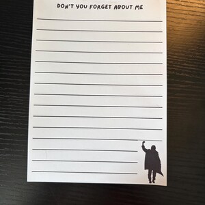 Op de afbeelding: Een wit rechthoekig notitieblok met zwarte horizontale lijnen. Bovenaan de pagina staat de tekst "DON'T YOU FORGET ABOUT ME" in een eenvoudig zwart lettertype. In de rechterbenedenhoek staat een zwart silhouet van een persoon met een vuist.