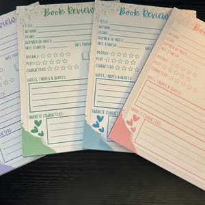 Pode incluir: Quatro blocos de notas coloridos para resenhas de livros. Cada bloco de notas apresenta seções para título, autor, gênero e classificações. Os blocos têm um design de borda de papel rasgado com corações e estrelas. O texto "Book Review" está no topo.