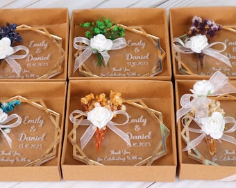 May include: Six small, square boxes each containing a hexagonal, clear glass ornament with a gold frame. Each ornament features a small bouquet of dried flowers and a personalized message with the names "Emily & Daniel" and a date.