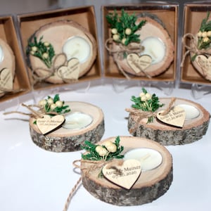 May include: Set of rustic wooden candle holders with small white candles. Each holder is decorated with a small sprig of greenery and a heart-shaped tag with the text "Ashton & Mainez 07.30.2020".