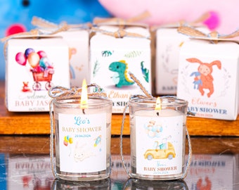 May include: Lit candles in clear glass holders with silver wire handles, labeled for baby showers. Each candle has a label with a baby-themed illustration and the text "Leo's BABY SHOWER 20.04.2024" or "Then's BABY SHOWER 20.04.2024". Small gift boxes with baby-themed illustrations are in the background.