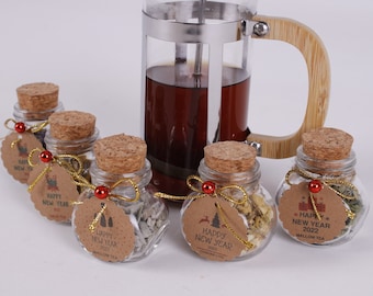 May include: Five small glass jars with cork lids filled with dried herbs and tied with twine and a small tag. The tags are brown and have the text "Happy New Year 2022" printed on them. The jars are arranged in a semi-circle on a white surface.