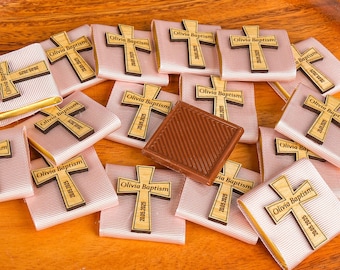 May include: A collection of favors for a baptism, featuring small, light pink wrapped squares topped with wooden crosses. Each cross is engraved with "Olivia Baptism" and the date "20.05.2025". A square of chocolate sits among the favors.