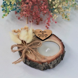 Op de afbeelding: Een houten hart met de tekst "Kevin & Danielle 9-12-21" is bevestigd aan een klein houten schijfje met een kaarshouder. Gedroogde witte bloemen zijn op het houten schijfje gerangschikt.
