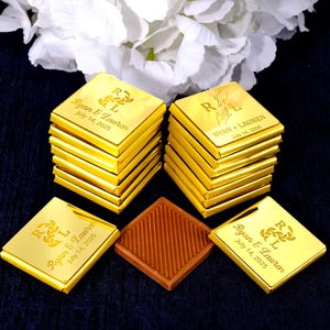 May include: Gold-wrapped chocolate squares, personalized with the names "Ryan & Lauren" and the date "July 14, 2025", are stacked and arranged on a dark blue surface. A single brown square with a textured pattern is also present. White flowers are in the background.