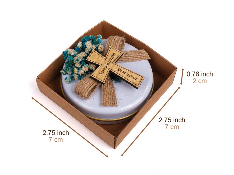 May include: A baptism favor in a brown box, measuring 2.75 inches (7 cm) square and 0.78 inches (2 cm) high. It features a wooden cross with the text "Tom's Baptism" and the date "25.07.2025", along with blue and white flowers.