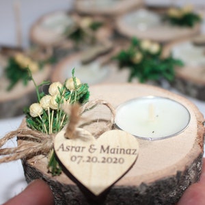 May include: Wooden candle holder with a heart-shaped tag that says "Asrar & Mainaz 07.20.2020". The holder is decorated with greenery and small white flowers.