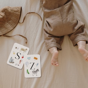 May include: Two white flash cards with colorful illustrations of objects and the letters "S" and "Z" on a white background. The cards are on a beige surface with a brown hat and a baby's feet.