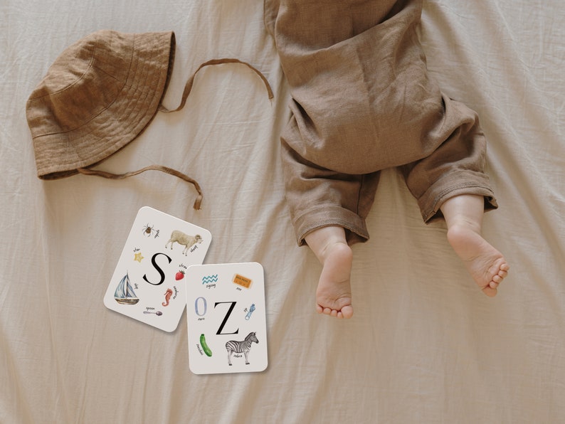 May include: Two white flash cards with colorful illustrations of objects and the letters "S" and "Z" on a white background. The cards are on a beige surface with a brown hat and a baby's feet.
