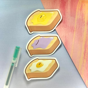 Może przedstawiać: Trzy ilustrowane naklejki z tostami z różnymi dodatkami. Pierwszy tost ma żółty dodatek masła, drugi fioletowy dodatek galaretki, a trzeci biały dodatek jajka.