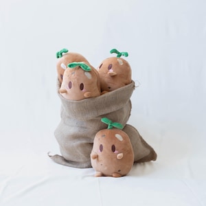 May include: A collection of plush potato toys with smiling faces and green leaf accents. The potatoes are in a burlap sack, with one standing in front. The toys are a light brown color with pink cheeks and small black eyes.