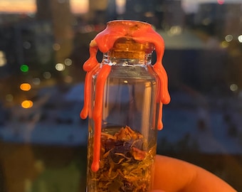 May include: A small glass bottle with a cork stopper is filled with dried herbs and flowers. The bottle is sealed with a pink wax seal that is dripping down the sides.