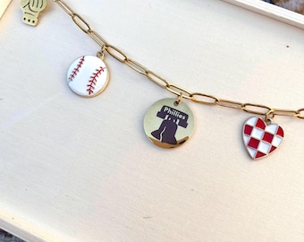 May include: A gold chain bracelet with charms featuring a baseball, a heart with a checkered pattern, a Liberty Bell, and a baseball glove. The charms are attached to a gold chain.
