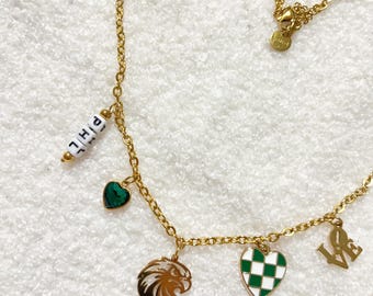 May include: Gold chain necklace with multiple charms, including a green heart, a green and white chequered heart, a gold eagle head, and a gold charm with the word "LOVE" on it. The necklace also has a white bead charm with the word "GIRL" on it.