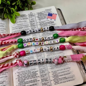 Könnte beinhalten: Offene Bibel mit Perlen-Lesezeichen mit den Wörtern "Psalm 23", "Gott bete zuerst", "Matt 19:26" und "Lob". Die Lesezeichen sind an bunten Stoffstreifen befestigt. Eine kleine amerikanische Flagge ist sichtbar.