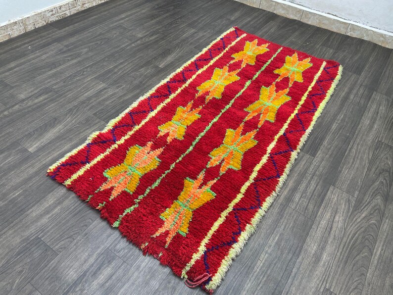 May include: A rectangular red rug with a repeating pattern of yellow and orange star-like shapes. The rug has a textured appearance with a yellow border and a dark wood floor background.