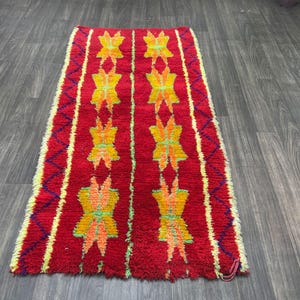 May include: A rectangular red rug with a repeating pattern of yellow and orange floral designs. The rug has a textured appearance and is bordered by yellow and purple zigzags. The rug is placed on a wooden floor.