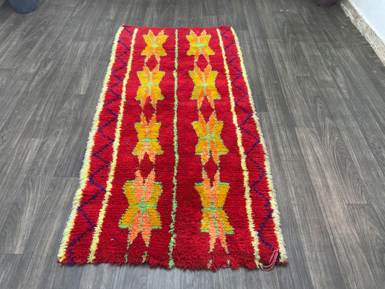 May include: A rectangular red rug with a repeating pattern of yellow and orange floral designs. The rug has a textured appearance and is bordered by yellow and purple zigzags. The rug is placed on a wooden floor.