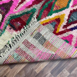 May include: A colorful Moroccan rug with a geometric pattern in shades of pink, red, green, yellow, and blue. The rug has a fringe border and is folded in half.