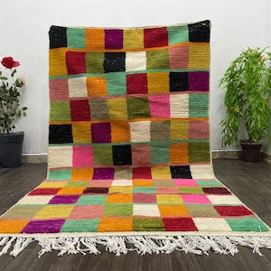 Peut inclure: Un tapis coloré tissé à la main avec un motif à carreaux dans des tons de rouge, vert, jaune, rose et blanc. Le tapis a une bordure à franges et est fait de laine.