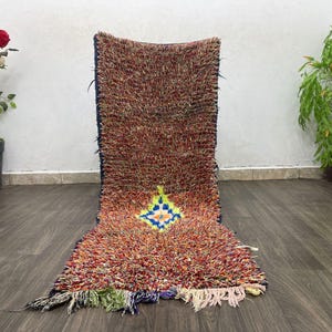 Puede incluir: Una alfombra larga y rectangular con una textura peluda. La alfombra es principalmente roja y naranja con un patrón geométrico azul y amarillo en el centro.