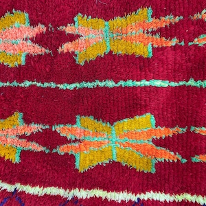 May include: Close-up of a red, textured rug with a repeating pattern of stylized floral shapes in yellow, orange, and pink, outlined in turquoise. Horizontal stripes in white and turquoise add contrast.