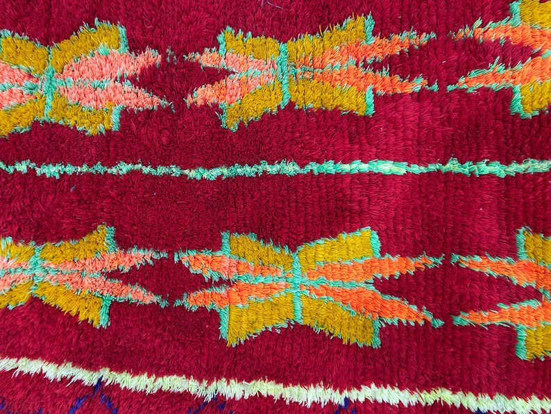 May include: Close-up of a red, textured rug with a repeating pattern of stylized floral shapes in yellow, orange, and pink, outlined in turquoise. Horizontal stripes in white and turquoise add contrast.