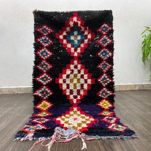 May include: A black, red, white and blue Moroccan rug with a geometric pattern. The rug is hand-woven and has a fringe on the bottom.