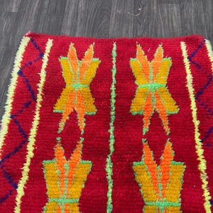 May include: A vibrant red rug with a repeating pattern of stylized floral designs in yellow, orange, and green. The rug has a textured surface and a yellow and blue geometric border. The rug is likely handmade.
