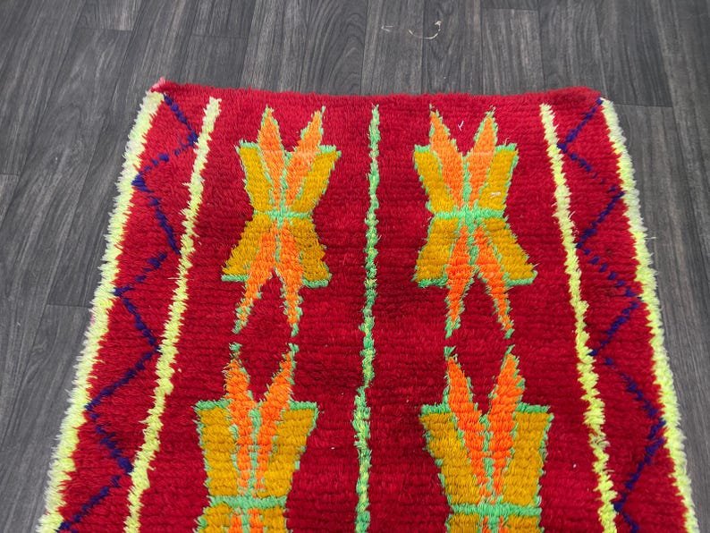 May include: A vibrant red rug with a repeating pattern of stylized floral designs in yellow, orange, and green. The rug has a textured surface and a yellow and blue geometric border. The rug is likely handmade.