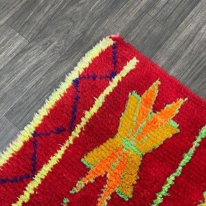 May include: Close-up of a vibrant red rug with a textured, shaggy pile. The rug features a bold design with yellow, orange, and green geometric shapes and stripes. The rug's edges are trimmed with a yellow border.