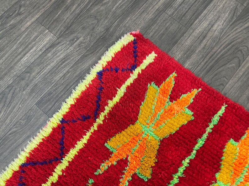 May include: Close-up of a vibrant red rug with a textured, shaggy pile. The rug features a bold design with yellow, orange, and green geometric shapes and stripes. The rug's edges are trimmed with a yellow border.