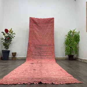 Puede incluir: Una alfombra larga y rectangular, tejida a mano, en tonos rosas y rojos. La alfombra presenta una superficie texturizada con un patrón geométrico en la parte inferior y líneas horizontales oscuras. La alfombra se exhibe sobre un suelo de madera oscura.