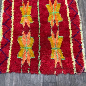 May include: A vibrant red rug with a repeating pattern of stylized floral motifs in yellow, orange, and green. The rug has a textured, woven appearance with a fringed edge and yellow and blue accents.