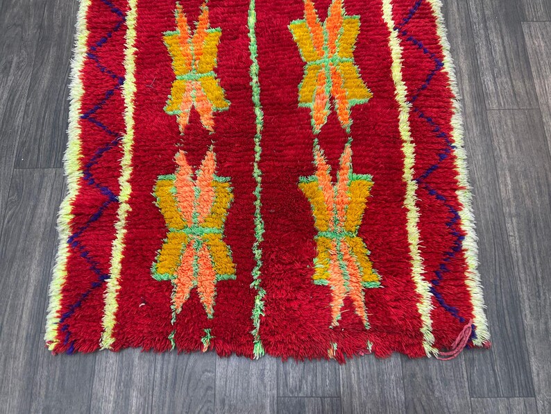 May include: A vibrant red rug with a repeating pattern of stylized floral motifs in yellow, orange, and green. The rug has a textured, woven appearance with a fringed edge and yellow and blue accents.