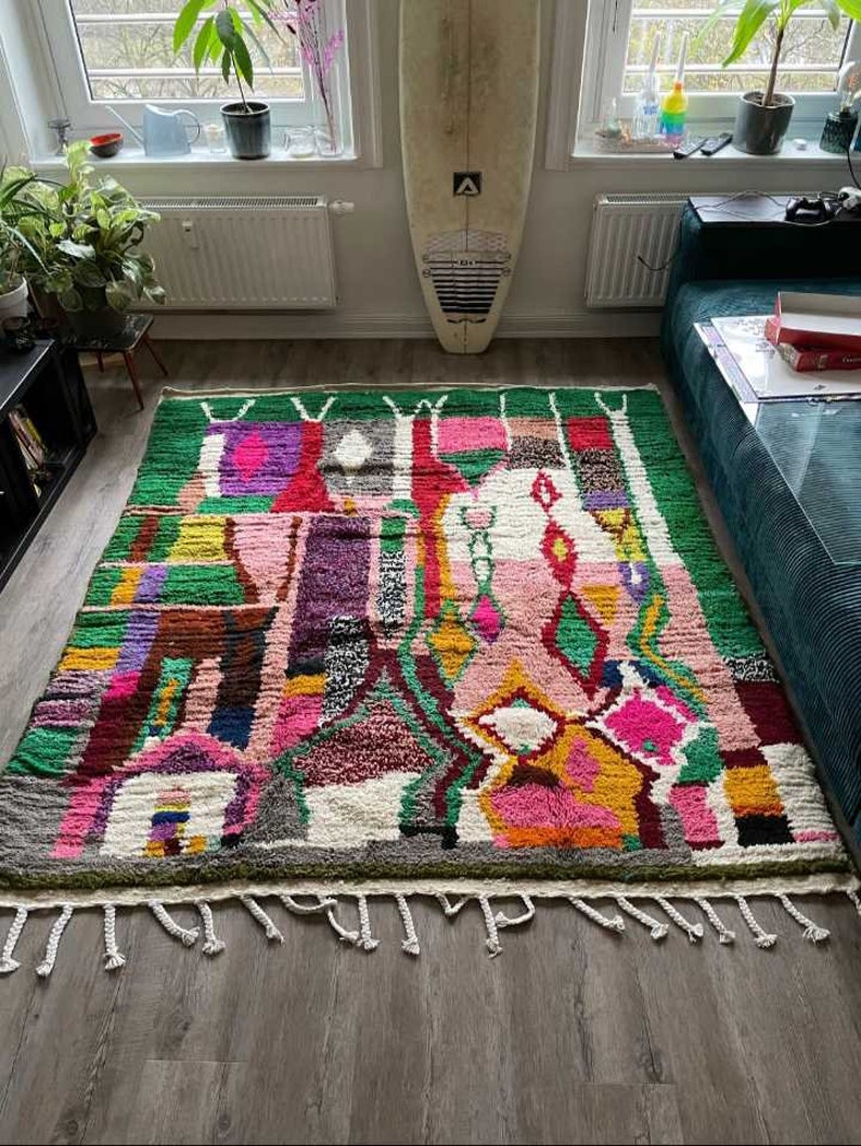 May include: A colorful, abstract rug with a green background and pink, yellow, and purple accents. The rug has a fringe border and is made of wool.