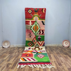 Op de afbeelding: Een kleurrijk Marokkaans tapijt met geometrische patronen in tinten rood, groen, roze en wit. Het tapijt heeft een franje rand en is gemaakt van wol.