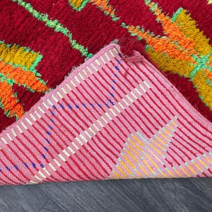 May include: A vibrant, hand-woven rug with a red background and colorful geometric patterns in orange, yellow, and green. The underside reveals a pink and white striped pattern. The rug's texture is visible, suggesting a soft, comfortable feel.
