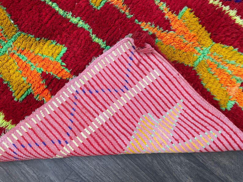 May include: A vibrant, hand-woven rug with a red background and colorful geometric patterns in orange, yellow, and green. The underside reveals a pink and white striped pattern. The rug's texture is visible, suggesting a soft, comfortable feel.