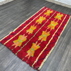May include: A rectangular red rug with a repeating pattern of yellow and orange star-like shapes. The rug has a textured appearance with yellow and blue accents along the edges. The rug is placed on a dark wood floor.