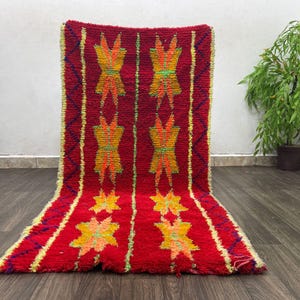May include: A vibrant red rug with a geometric pattern. The rug features repeating yellow and orange star-like designs and vertical stripes. The rug is made of a thick, textured material and is displayed on a wooden floor.