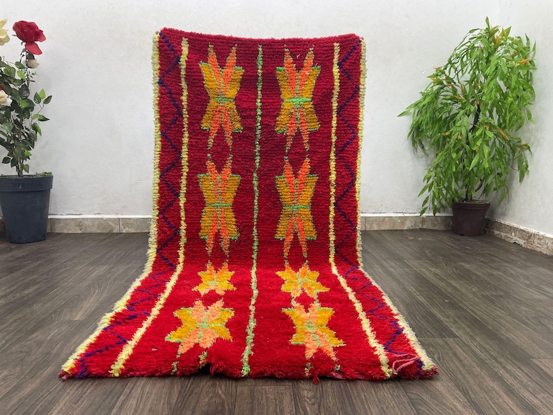 May include: A vibrant red rug with a geometric pattern. The rug features repeating yellow and orange star-like designs and vertical stripes. The rug is made of a thick, textured material and is displayed on a wooden floor.