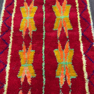 May include: A vibrant red rug with a repeating pattern of yellow, orange, and green geometric shapes. The rug has a textured appearance and a border with blue and yellow diamond shapes. The design is symmetrical and eye-catching.
