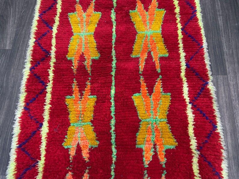May include: A vibrant red rug with a repeating pattern of yellow, orange, and green geometric shapes. The rug has a textured appearance and a border with blue and yellow diamond shapes. The design is symmetrical and eye-catching.