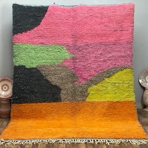 May include: A colorful, abstract rug with a geometric design. The rug features a vibrant palette of black, pink, green, brown, yellow, and orange. The rug has a shaggy texture and a fringe along the bottom edge. This rug is suitable for a living room or bedroom.