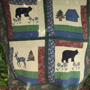 May include: A beige and green quilt with a patchwork design featuring black bears, deer, trees, and houses. The quilt has a blue and red border.